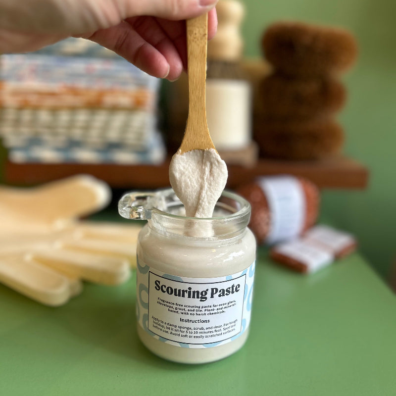 A hand holds a bamboo spoon while it drizzles scouring scub into the jar. The fragrance free scrub is thick and ready to take on tough kitchen messes. 