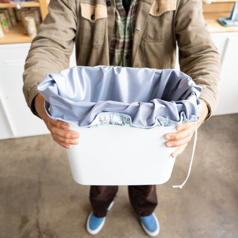 A person holding a small trash bin with a grey drawstring reusable pail liner in it . 