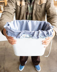 A person holding a small trash bin with a grey drawstring reusable pail liner in it . 