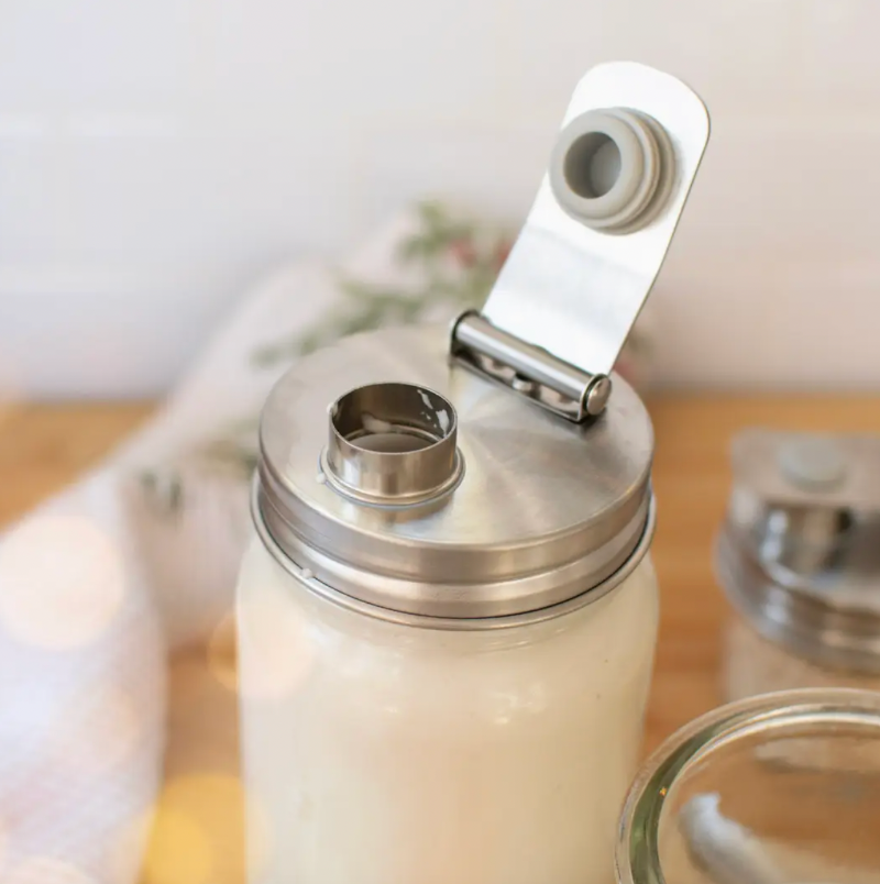 A stainless steel mason jar lid with a flip-up spout, shown on a mason jar filled with a white beverage. The jar is set on a wooden surface with a soft-focus background.
