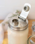 A stainless steel mason jar lid with a flip-up spout, shown on a mason jar filled with a white beverage. The jar is set on a wooden surface with a soft-focus background.