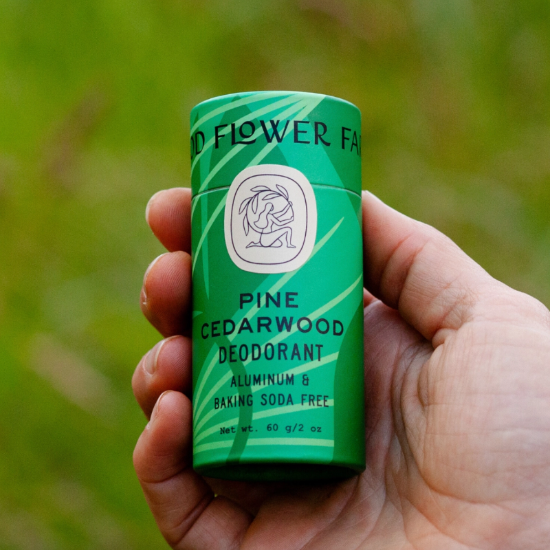 A hand holds a green cylindrical Good Flower Farm Deodorant Stick, which is aluminum and baking soda free, against a blurred green background.