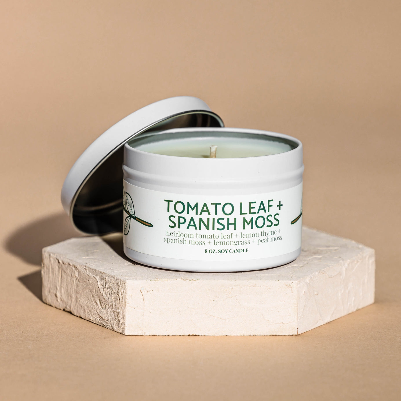 Candle in a container labeled 'Tomato Leaf + Spanish Moss' on a beige  background