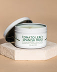 Candle in a container labeled 'Tomato Leaf + Spanish Moss' on a beige  background
