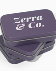 Three stacked square metal tins in a muted purple color, with the top tin displaying the white text 'Zerra & Co.' on its lid, set against a clean white background.