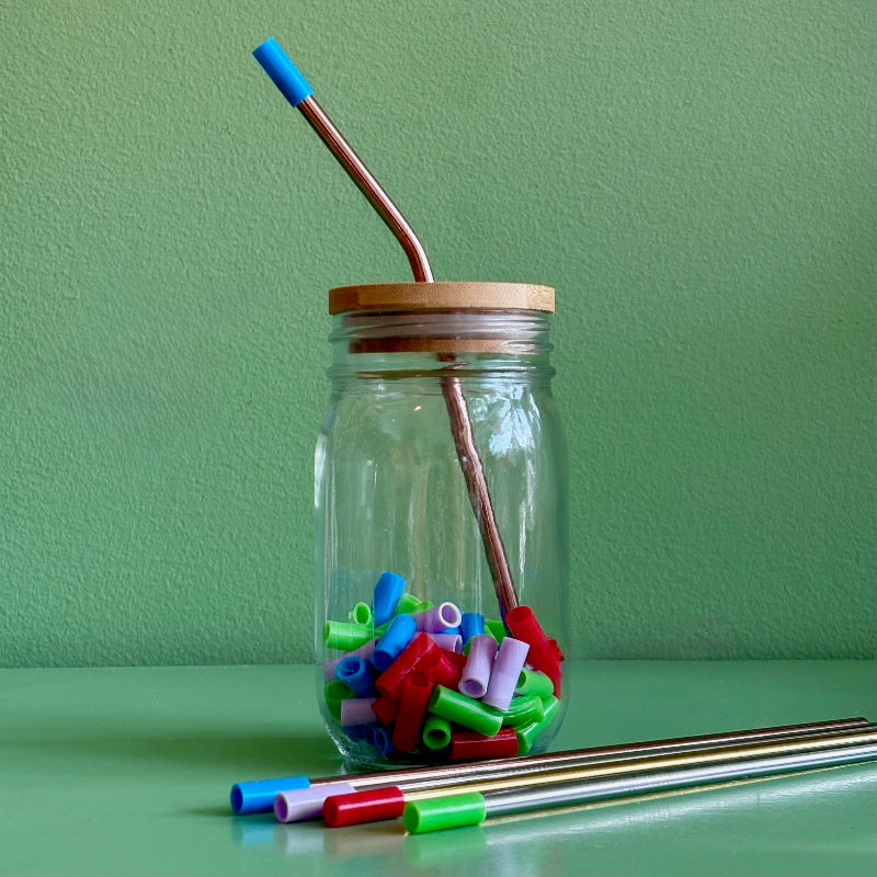 A reused mason jar with a bamboo straw lid and a rose gold metal straw with a blue silicone straw tip on it.  The jar is full of multi-colored straw tips and sits on a green background.