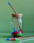 A reused mason jar with a bamboo straw lid and a rose gold metal straw with a blue silicone straw tip on it.  The jar is full of multi-colored straw tips and sits on a green background.