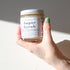 A hand with green-painted nails holds a Shade Tree Naturals Sugar Scrub jar under bright light, displaying some of the gently exfoliating sugar scrub on their hand.