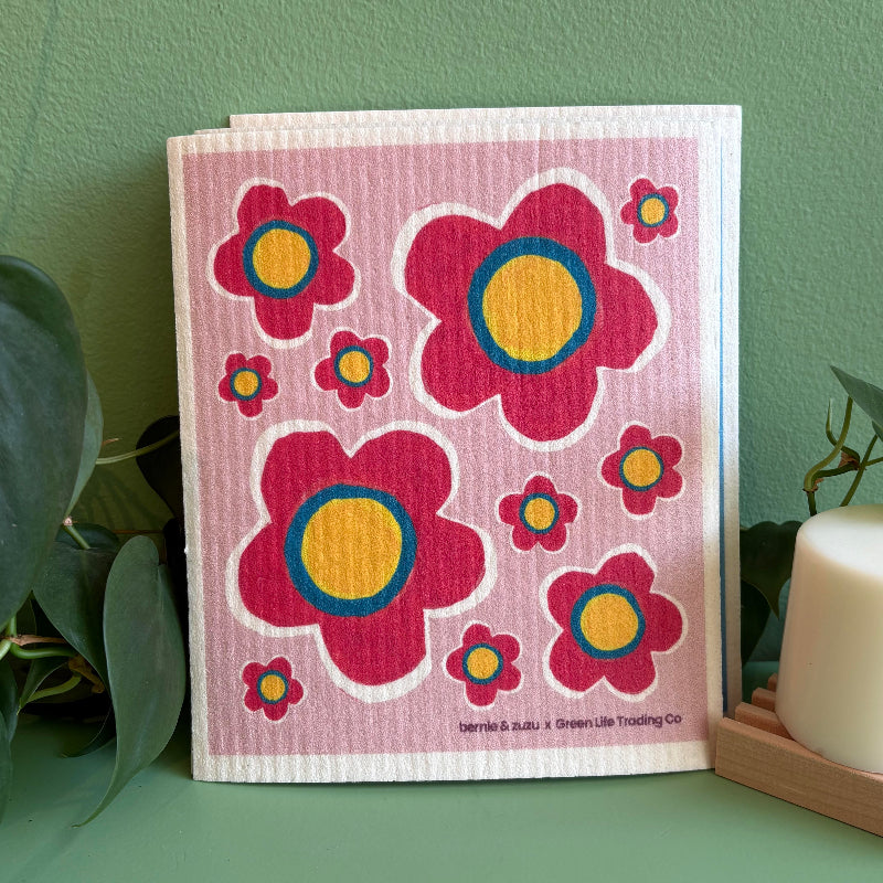 A pink Swedish dishcloth with a pink flower design on it by local Madison Artist Bernie & Zuzu. It is sitting on a green background by a plant and solid dish puck.