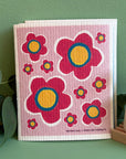 A pink Swedish dishcloth with a pink flower design on it by local Madison Artist Bernie & Zuzu. It is sitting on a green background by a plant and solid dish puck.