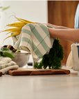 Person holding a green and white checkered towel over vegetables on a wooden cutting board in a kitchen.