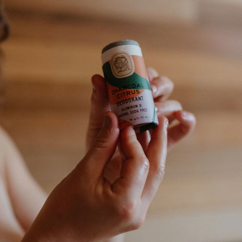 Someone holds a Good Flower Farm Deodorant Stick with a beige, green, and peach-colored label in their hands against a softly blurred background.