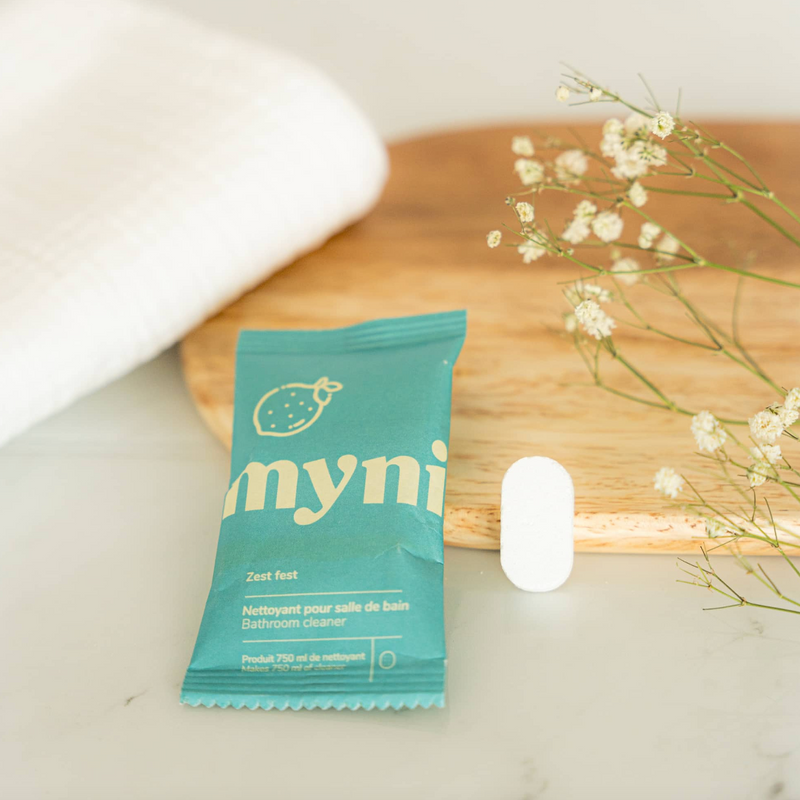 A teal Myni Bathroom Cleaning Tab packet rests on a white surface near a matching cleaning tablet, a wooden tray, a white towel, and small white flowers.