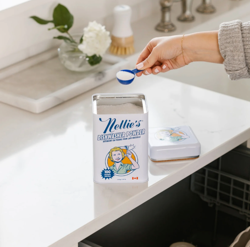 Person measuring Nellie's Dishwasher Powder with a scoop in a kitchen setting
