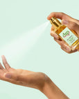 hand holding glass bottle of hair and body dry oil spraying mist over other hand on green background. 