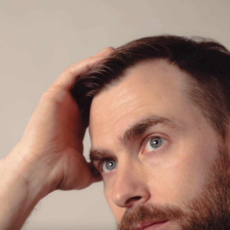 Man with a beard and blue eyes looking to the side with one hand on his head against a neutral background