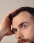 Man with a beard and blue eyes looking to the side with one hand on his head against a neutral background