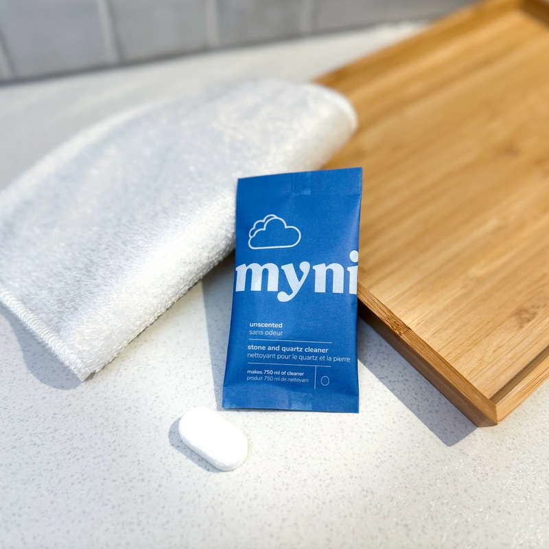 A blue Myni Stone & Quartz Cleaning Tab packet rests on a white countertop near a folded white towel, a white oval tablet, and a wooden cutting board.