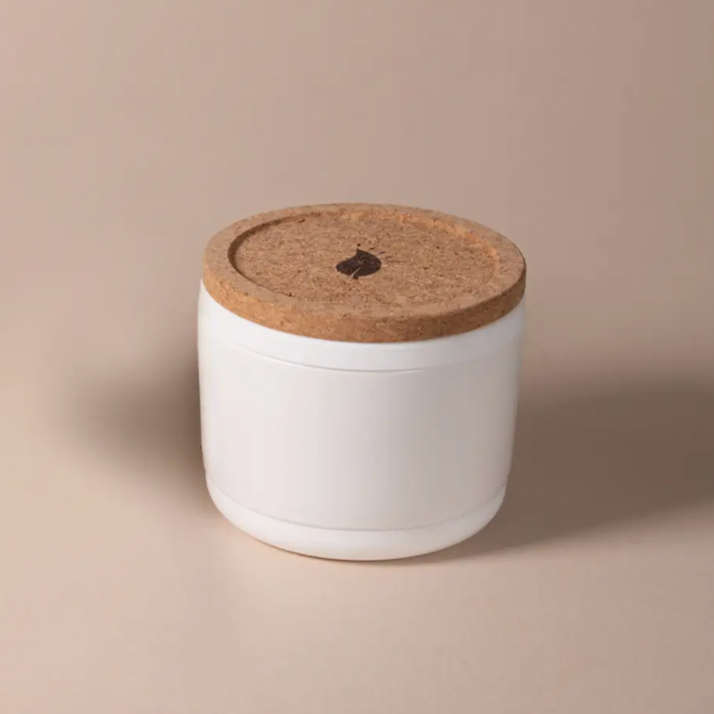 The Arbor Made Refillable Candle Jar by Arbor Made is a small white ceramic container with a round cork lid featuring a Leaf symbol and a silicone bottom—ideal for use as refillable candle jars with your favorite wax refills.