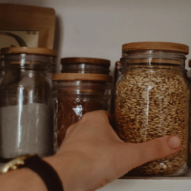 A hand reaches for a mason jar being reused to hold bulk goods on a shelf with other mason jars holding bulk goods. All the jars have bamboo lids. 