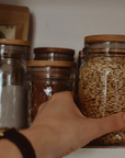 A hand reaches for a mason jar being reused to hold bulk goods on a shelf with other mason jars holding bulk goods. All the jars have bamboo lids. 