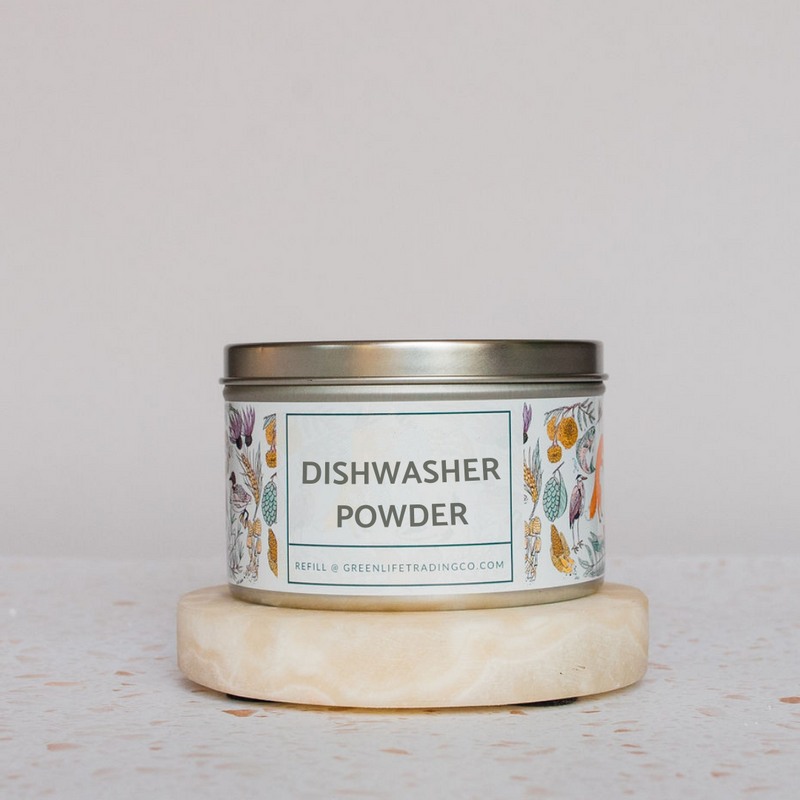 A metal tin labeled Nellies Dishwasher Powder, made with a biodegradable plant-based formula, sits on a round stone slab. The white label features botanical illustrations and greenlifetradingco.com below the main text.