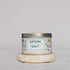 A tin of Salt Works Epsom Salt, labeled pharmaceutical-grade and decorated with botanical illustrations, sits on a round stone coaster against a light background, evoking classic bath products.