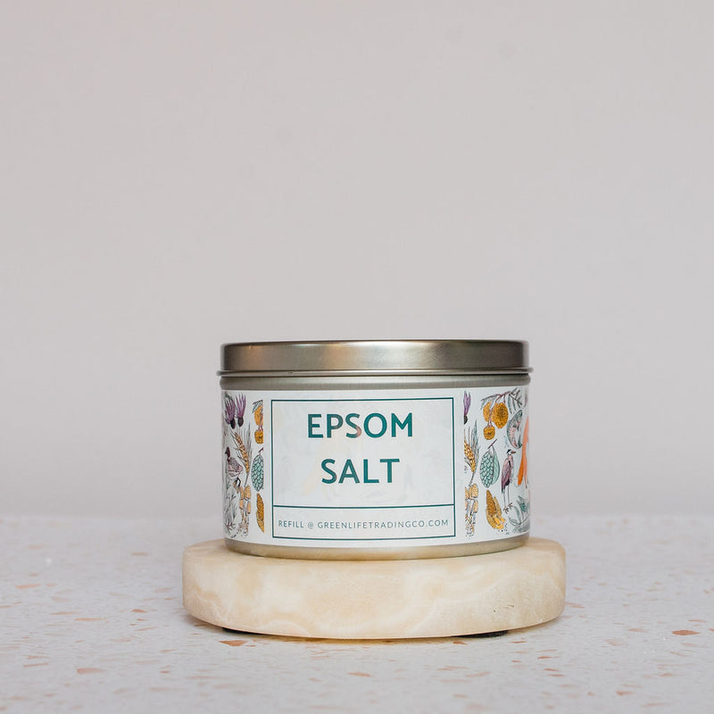 A tin of Salt Works Epsom Salt, labeled pharmaceutical-grade and decorated with botanical illustrations, sits on a round stone coaster against a light background, evoking classic bath products.