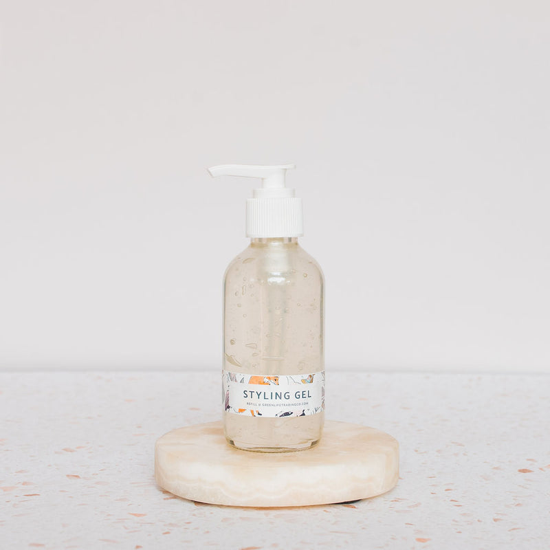 A clear pump bottle labeled Carina Organics Hair Styling Gel rests on a round, light stone with a white background. This alcohol-free gel defines curls while keeping a natural look.