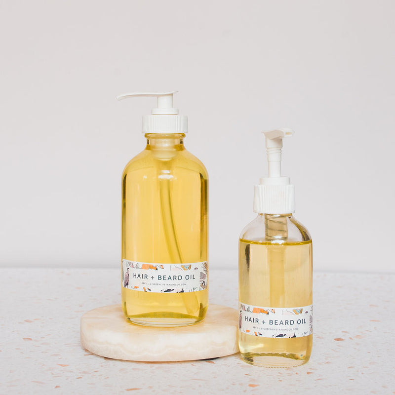 Two clear glass pump bottles of Green Life Trading Co. Hair, Beard & Body Oil, enriched with almond oil, rest on a round stone platform against a light, neutral background.