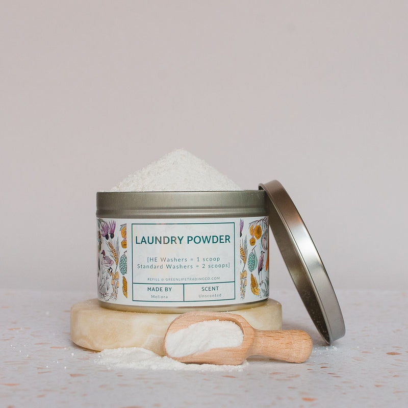 A tin of Meliora Laundry Powder with a floral label sits on a stone slab. Its lid leans to the side, and a wooden scoop filled with MADE SAFE Certified powder rests in front of the container.