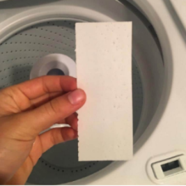 A hand holds a white plantbased Laundry Detergent Strip in front of an open washing machine drum.