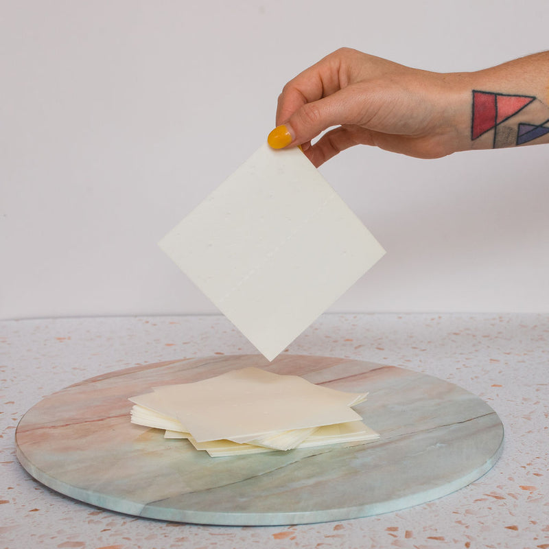 A hand with yellow nail polish and geometric tattoos holds a square sheet of TruEarth Laundry Detergent Strips above a stack of similar sheets on a round, marble-patterned surface.