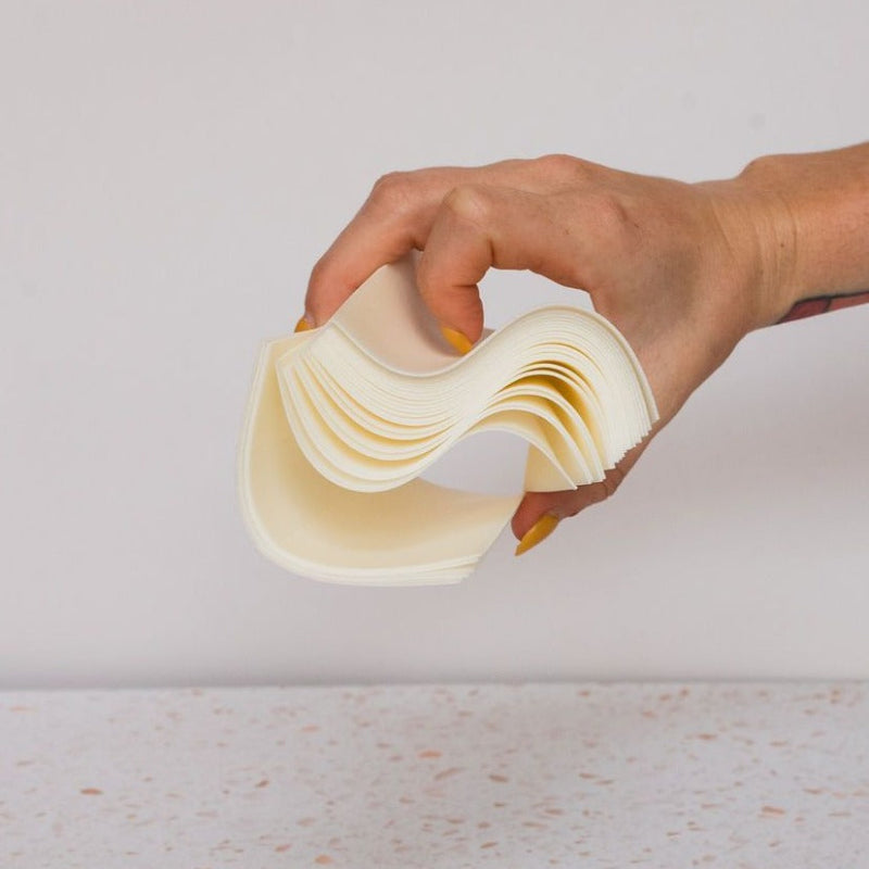 A hand with yellow-painted nails bends a stack of Plant based Laundry Detergent Strips against a plain, light background.