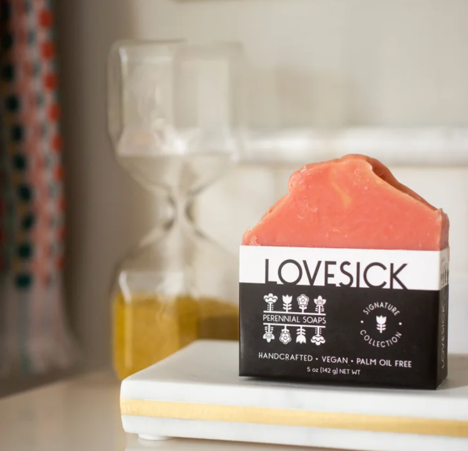 A Bar Soaps product by Perennial Soaps, vegan and handcrafted in Wisconsin without palm oil, sits on a white tray near an hourglass of yellow sand; its pink packaging highlights these natural qualities.
