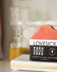 A Bar Soaps product by Perennial Soaps, vegan and handcrafted in Wisconsin without palm oil, sits on a white tray near an hourglass of yellow sand; its pink packaging highlights these natural qualities.