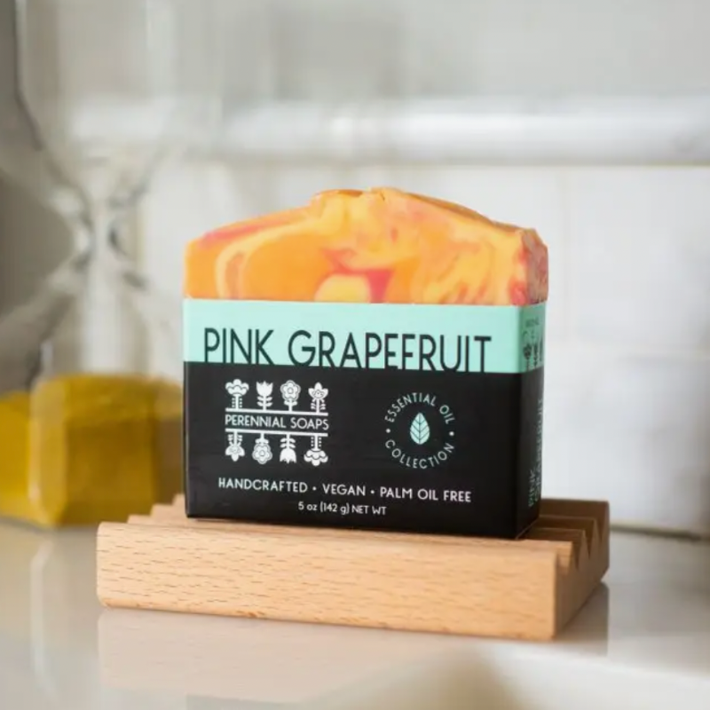 A vibrant bar of Perennial Soaps Bar Soaps, handcrafted in Wisconsin with orange and pink swirls and a turquoise label, highlights its vegan and palm oil free features. A blurred yellow soap sits in the background.