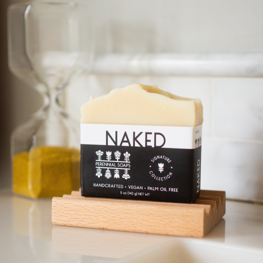 A Perennial Soaps Bar Soap rests on a wooden dish with an hourglass behind it. This handcrafted, vegan soap from Wisconsin is palm oil-free and features eco-friendly packaging.