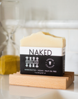 A Perennial Soaps Bar Soap rests on a wooden dish with an hourglass behind it. This handcrafted, vegan soap from Wisconsin is palm oil-free and features eco-friendly packaging.