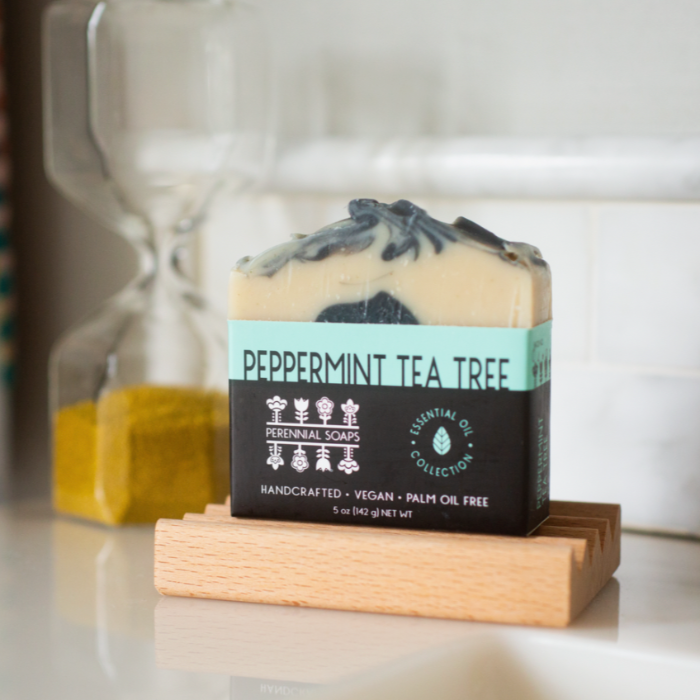 A bar of Perennial Soaps Bar Soap rests on a wooden dish near an hourglass and tiled wall. Handcrafted, vegan, and palm oil free, this soap is perfect for those who love handmade soaps.