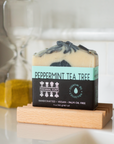 A bar of Perennial Soaps Bar Soap rests on a wooden dish near an hourglass and tiled wall. Handcrafted, vegan, and palm oil free, this soap is perfect for those who love handmade soaps.