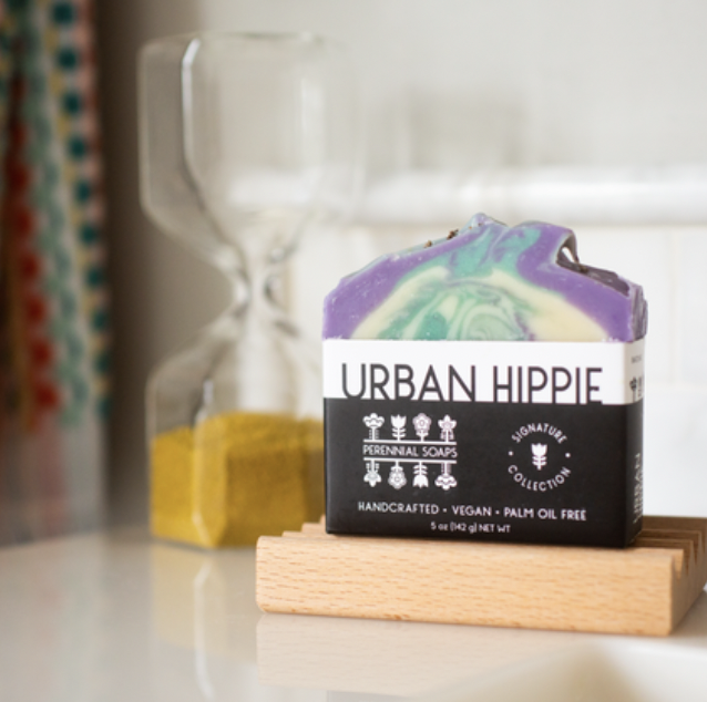 A colorful bar of Perennial Soaps Bar Soap rests on a wooden dish, with an hourglass of yellow sand behind it and a patterned curtain to the left, showcasing this vibrant, Wisconsin-made product.