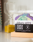 A colorful bar of Perennial Soaps Bar Soap rests on a wooden dish, with an hourglass of yellow sand behind it and a patterned curtain to the left, showcasing this vibrant, Wisconsin-made product.