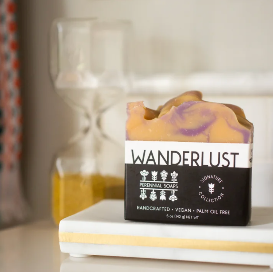 A Perennial Soaps Bar Soap sits on a white tray, its vegan tan and purple swirls on display. In the blurred background, a yellow-sand hourglass can be seen.