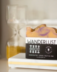 A Perennial Soaps Bar Soap sits on a white tray, its vegan tan and purple swirls on display. In the blurred background, a yellow-sand hourglass can be seen.