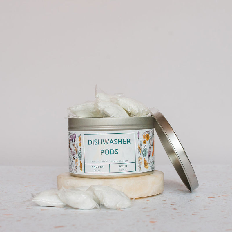 A Dropps Lemon Dish Pods tin sits on a round stone slab, its lid set aside. Several natural dish detergent pods are inside the tin, with a few Lemon Dish Pods placed in front.