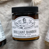 A brown jar labeled Larsons General Brilliant Bamboo Exfoliating Face Wash with a black lid sits on beige fabric. The label highlights bamboo powder, product details, and features a woman in sunglasses illustration.