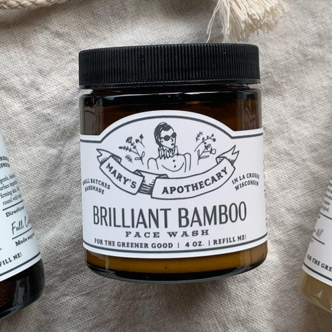 A brown jar labeled Larsons General Brilliant Bamboo Exfoliating Face Wash with a black lid sits on beige fabric. The label highlights bamboo powder, product details, and features a woman in sunglasses illustration.