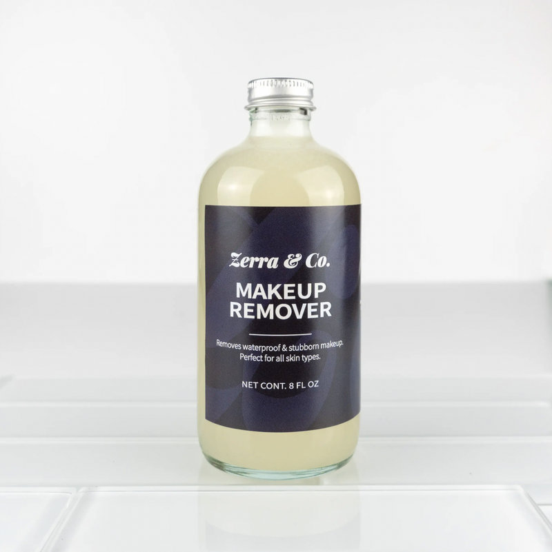 A clear glass 8 fl oz Zerra & Co. Makeup Remover sits on white tiles, filled with a light yellow liquid thats gentle on sensitive skin and proven to remove waterproof mascara and all types of makeup.