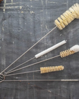 A collection of five natural fiber bottle brushes with metal handles laid out on a gray surface.